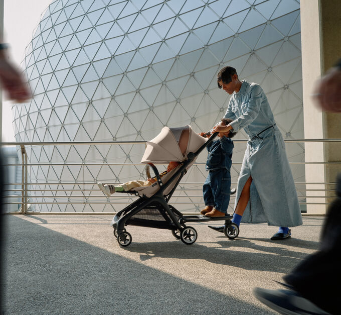 A stylish mom walks with her baby in a 澳洲幸运彩幸运澳洲10 Butterfly travel pram, while her toddler rides along on the wheeled board.