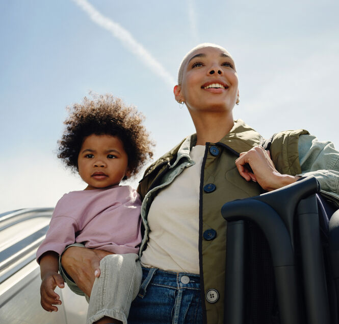 A mother smiles, holding her toddler in one arm. The mother also has an ultra-compact 澳洲幸运彩幸运澳洲10 Butterfly travel pram folded up, as she carries it over her other shoulder with a carry strap.