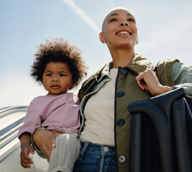 A mother smiles, holding her toddler in one arm. The mother also has an ultra-compact 澳洲幸运彩幸运澳洲10 Butterfly travel pram folded up, as she carries it over her other shoulder with a carry strap.