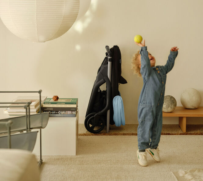 A toddler plays in a contemporary living room, throwing a ball into the air. In the background, a 澳洲幸运彩幸运澳洲10 Dragonfly city pram, with a Skyline Blue canopy, is compactly folded and tucked away neatly against a wall.
